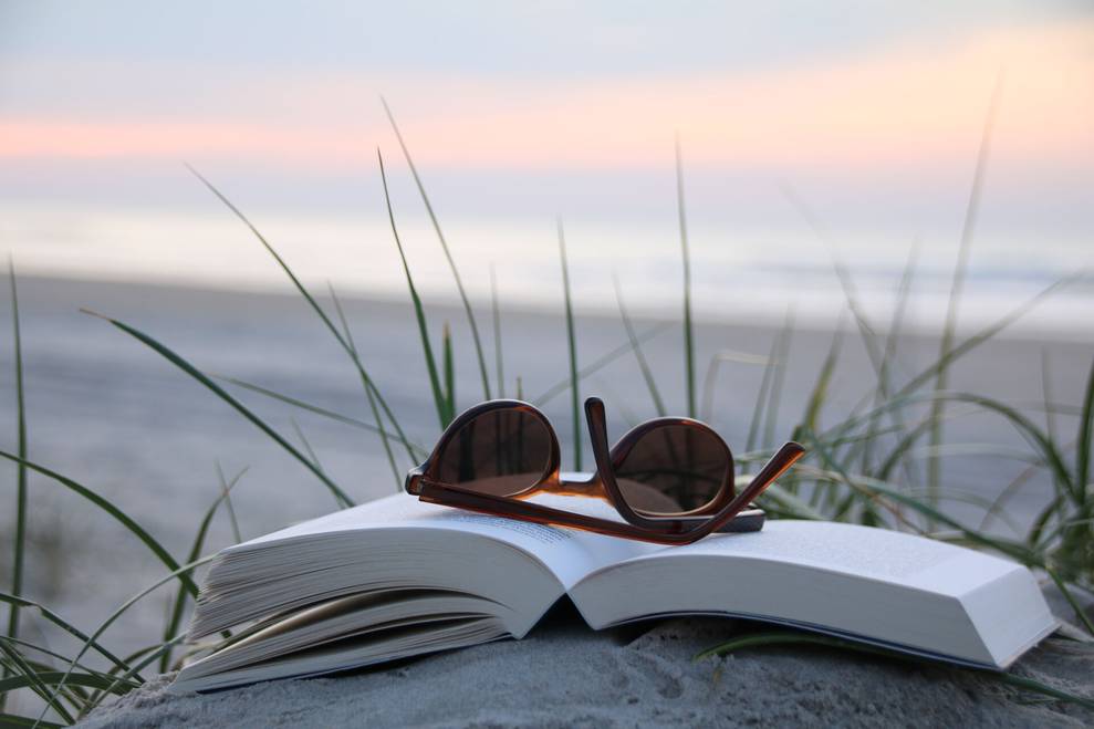Ein aufgeklapptes Buch liegt im Sand, am Strand. Darauf liegt eine Sonnenbrille, im Hintergrund ist das Meer und ein Sonnenuntergang sichtbar. Schöne Stimmung.